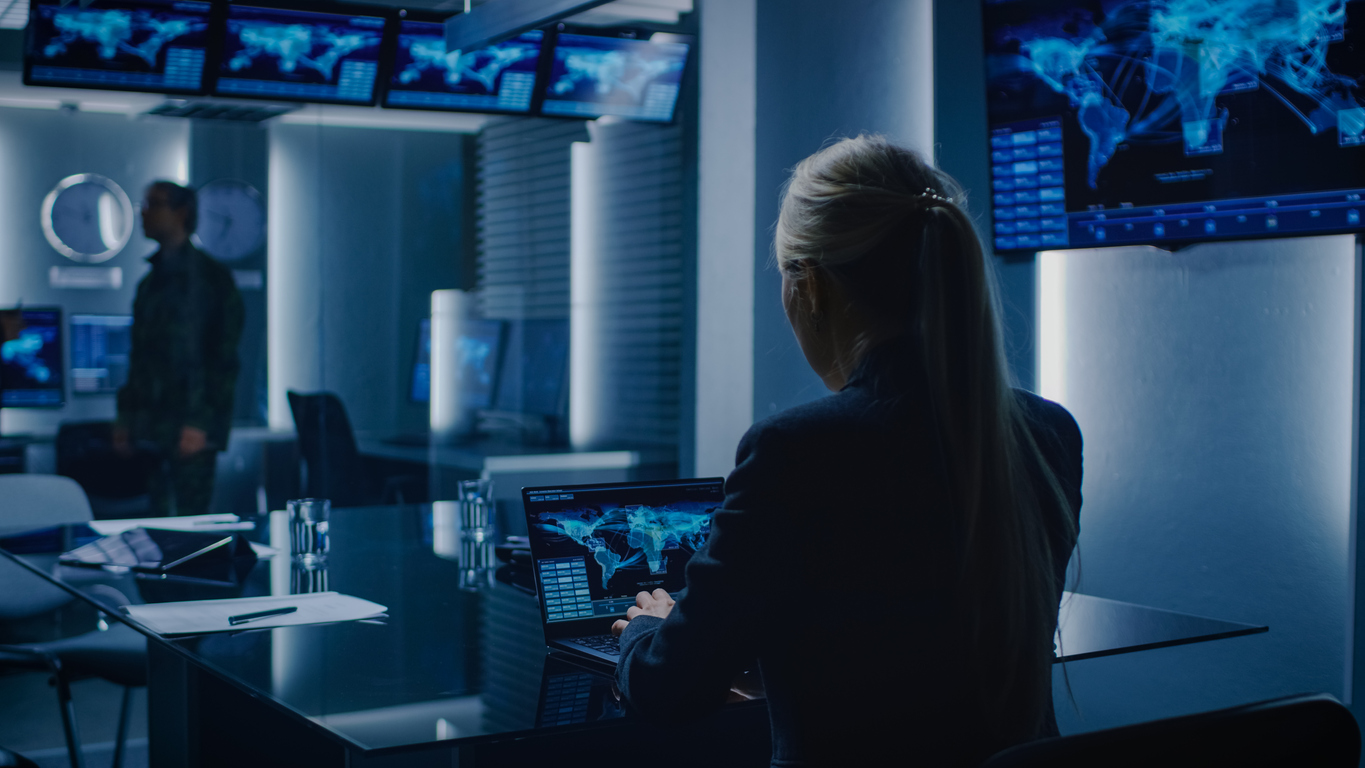 Female Special Agent Works on a Laptop in the Background Special Agent in Charge Talks To a Military Man in the Monitoring Room. In the Background Busy System Control Center with Monitors Showing Data Flow.